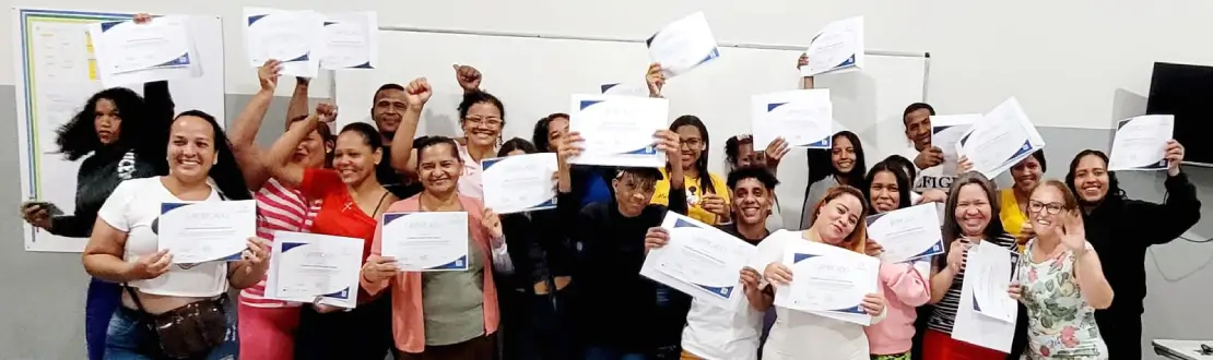 Foto de um grupo diverso de estudantes em uma sala de aula. Estão em pé, bem próximos, e seguram nas mãos os diplomas de conclusão do curso. Todos sorriem.
