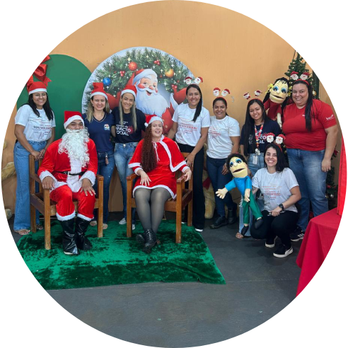 Foto dos voluntários caracterizados para uma ação de natal, posando ao lado do papai noel, em frente a uma decoração de natal.