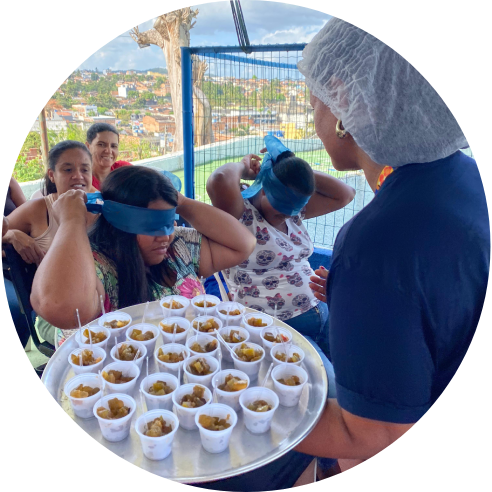 Foto de uma voluntária oferecendo copinhos com comida para crianças vendadas.