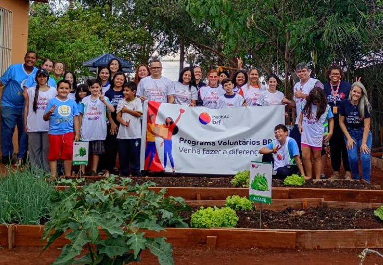 Foto de um grupo de voluntárias e voluntários atrás de vários canteiros de hortas. Alguns deles seguram um banner com o logo do Instituto.