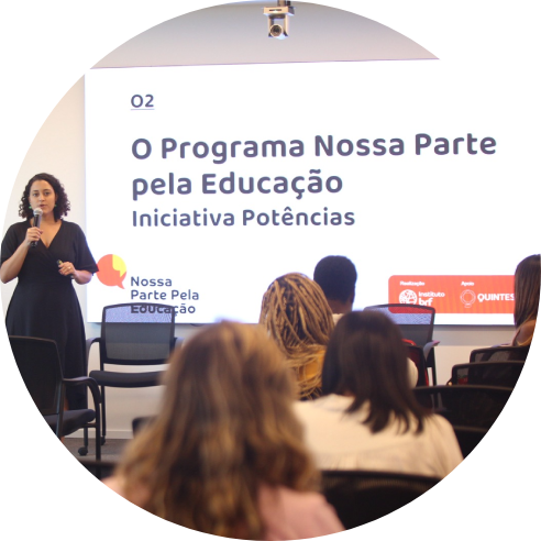 Foto de Jennifer França, da equipe do Instituto BRF, em pé, com um microfone na mão falando para um grupo de pessoas sentadas. Atrás dela, há um painel com o texto "O Programa Nossa Parte pela Educação. Iniciativa Potências."