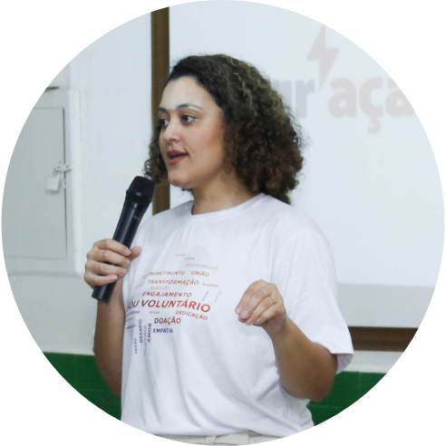 Foto de Jennifer França, da equipe do Instituto BRF, em pé, com um microfone na mão direita. Ela usa uma camiseta branca com uma nuvem de palavras referentes ao voluntariado.