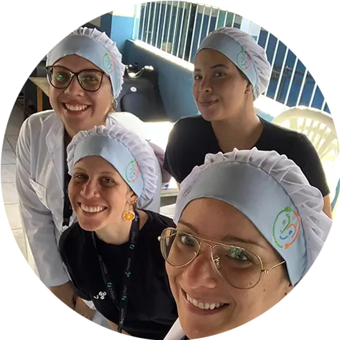 Foto em estilo selfie de um grupo de quatro mulheres em pé, juntinhas, usando tocas com o logo do programa Alimentação Escolar Inteligente. Todas sorriem.