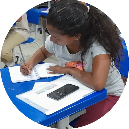 Foto em close de uma aluna sentada em uma sala de aula. Ela faz anotações em um caderno.