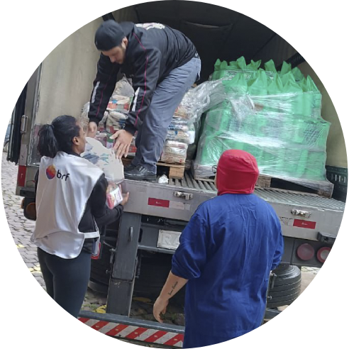 Foto de uma voluntária entregando itens arrecadados para um voluntário que coloca as doações em um caminhão. À direita deles, há outra pessoa voluntária.