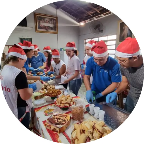 Foto de um grupo de voluntários e voluntárias do Instituto BRF colocando alimentos e bebidas em uma mesa. Todos usam gorros de Natal na cabeça.