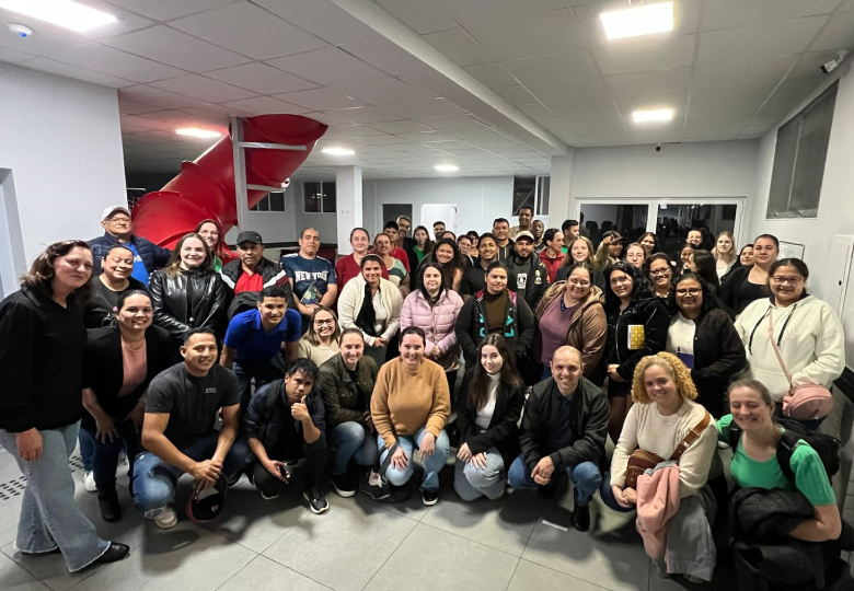 Foto de um grupo de pessoas, bem juntinhas, sendo que algumas estão sentadas e outras em pé, em uma sala.