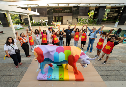 Foto de um grupo de pessoas e crianças de mãos dadas e os braços levantados ao redor da estátua de vaca customizada.