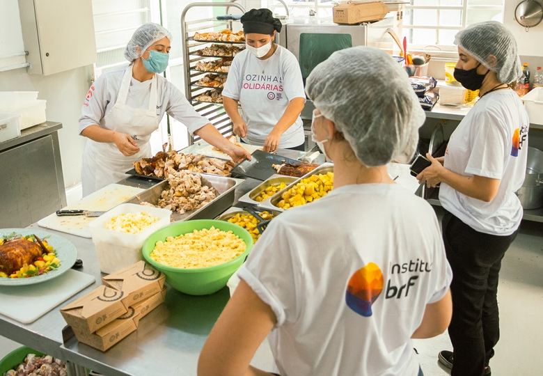 – Foto de integrantes das Cozinhas Solidárias e do Instituto MBRF em uma cozinha industrial, organizando e preparando alimentos em uma bancada com diversos recipientes e utensílios.