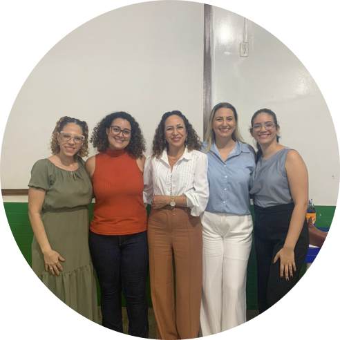 A imagem mostra cinco mulheres vestidas de maneira formal lado a lado, sorrindo, em uma sala de aula.