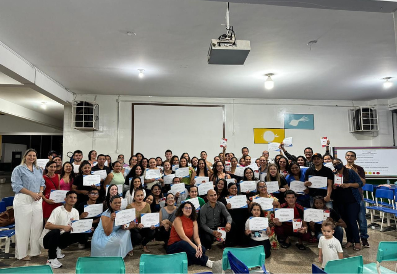 Foto de um grupo grande de pessoas em uma sala, segurando certificados e posando para uma foto em grupo. A maioria está sorrindo, celebrando a conclusão do curso.