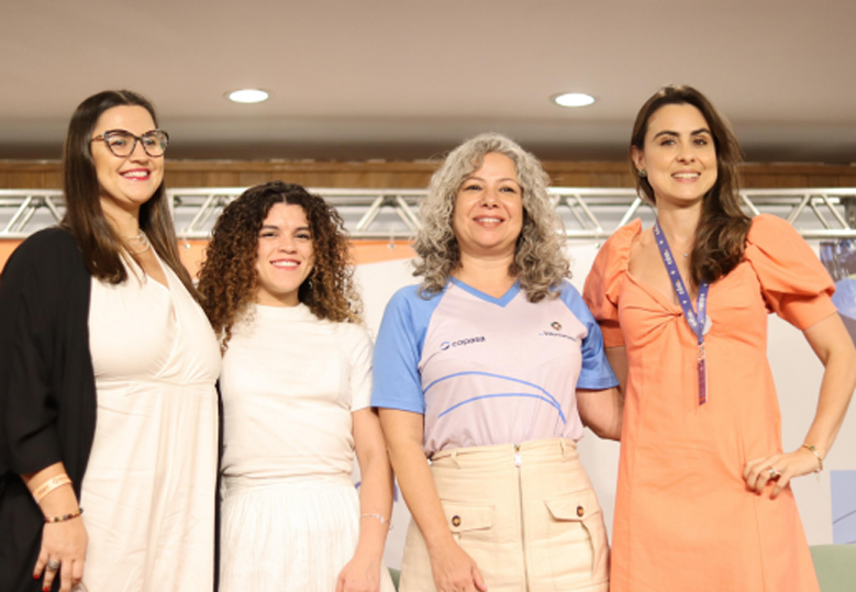 A imagem mostra quatro mulheres sorrindo e posando para a foto em um ambiente do evento. Elas estão lado a lado, com roupas casuais e formais. Ao fundo, há um banner com logotipos e uma estrutura metálica típica de eventos.