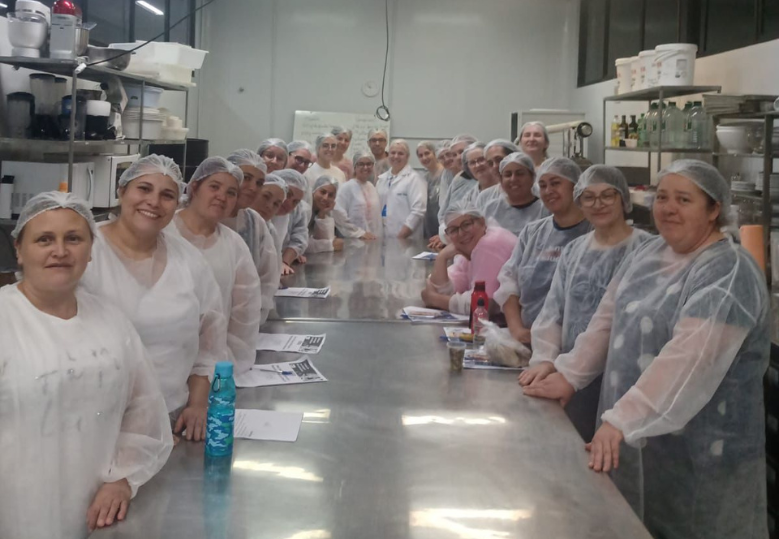 A imagem mostra um grupo grande de pessoas, em sua maioria merendeiras da prefeitura, vestindo toucas e jalecos de proteção em uma cozinha industrial. Elas estão reunidas em volta de uma longa mesa de metal, olhando para a câmera e sorrindo.