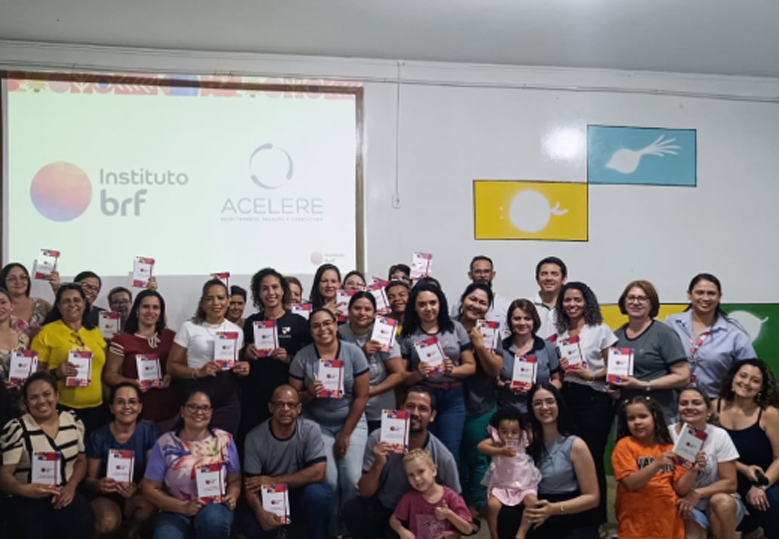 A imagem mostra um grupo grande de pessoas, em sua maioria mulheres, sorrindo e posando juntas, segurando cadernos com o logo do “Instituto BRF”. Ao fundo, há uma projeção com os logos do "Instituto BRF" e do "Acelere".