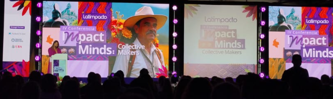 Foto do palco do evento com a mestre de cerimônias na frente de um púlpito. No telão, há imagem de um homem com chapéu e o nome do evento.
