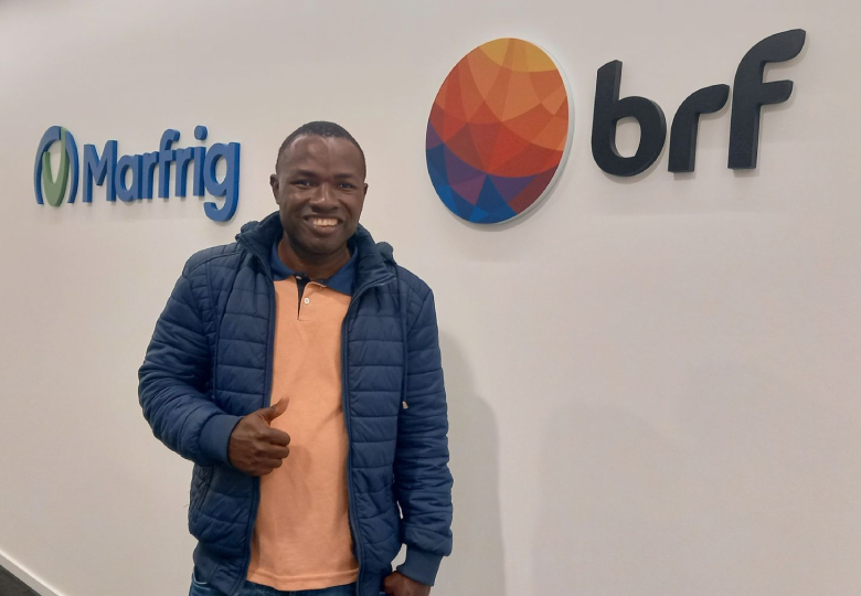 Foto de uma pessoa em frente a uma parede branca com os logotipos da Marfrig e da BRF, posando com uma das mãos levantada.