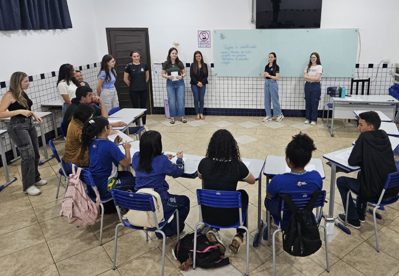 Foto de uma atividade em sala de aula, com estudantes sentados em círculo, e as voluntárias estão à frente, próximo ao quadro.