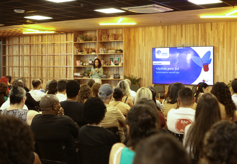 Foto do evento +juntos pelo sul em auditório, com público sentado enquanto a Diretora Raquel Ogando apresenta o evento.