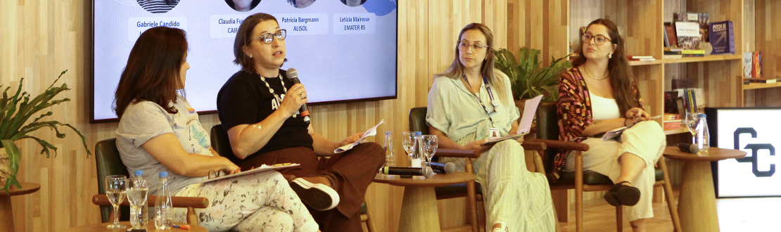 Foto de um painel com 4 participantes sentado em poltronas no palco, cada um com microfone. Tela ao fundo exibe o nome das pessoas convidadas.