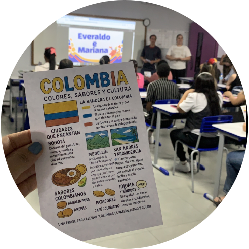 Foto com destaque para um folder com informações da Colômbia em espanhol e ao fundo a apresentação de um grupo de estudantes.