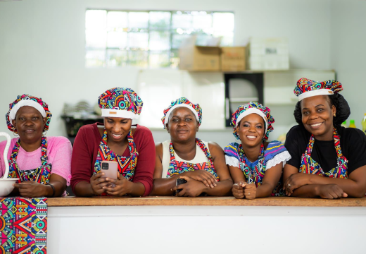 Foto de mulheres de pele negra com aventais e toucas coloridas em uma cozinha. Elas estão com os braços apoiados em cima de um balcão, lado a lado e sorriem.