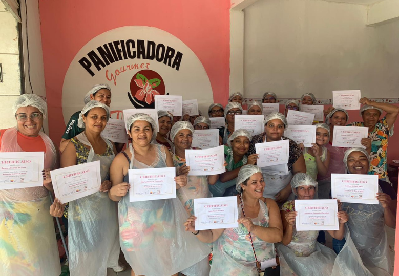 Um grupo de mulheres segura certificados, usando aventais plásticos e toucas. Ao fundo, um letreiro com "Panificadora Gourmet".