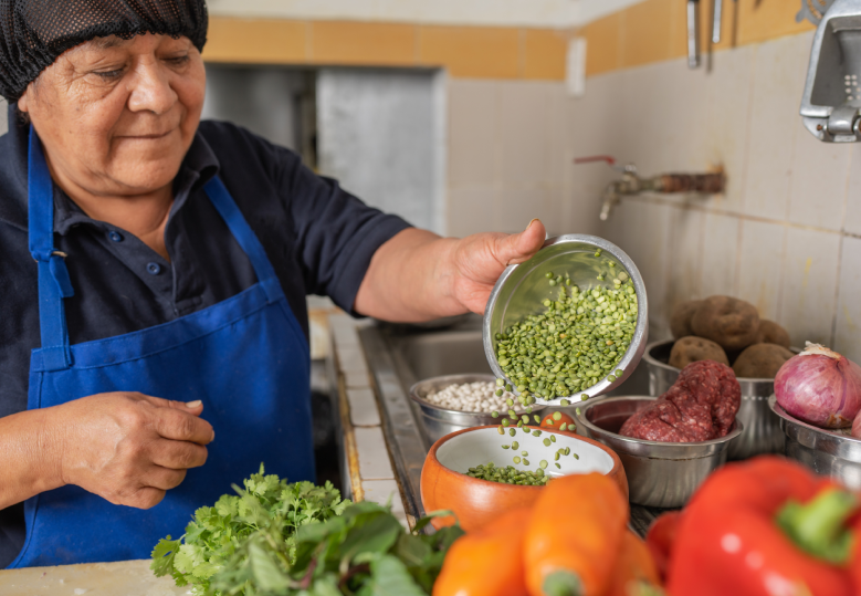 A imagem mostra uma mulher usando touca e avental azul, despejando grãos verdes de uma tigela em um recipiente na cozinha. Na bancada, há ingredientes como pimentões, ervas frescas, batatas, cebolas e carne.