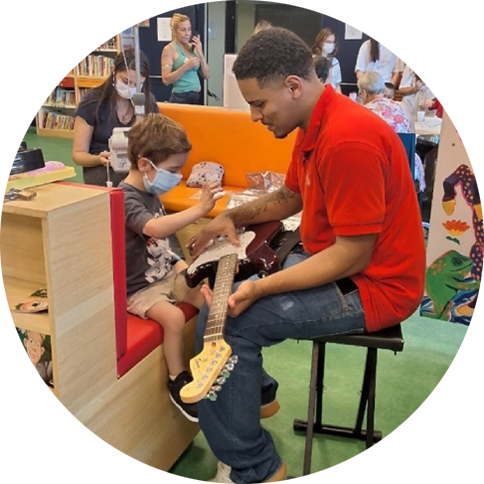 Foto de um voluntário do Instituto segurando uma guitarra para que um menino possa mexer em suas cordas.