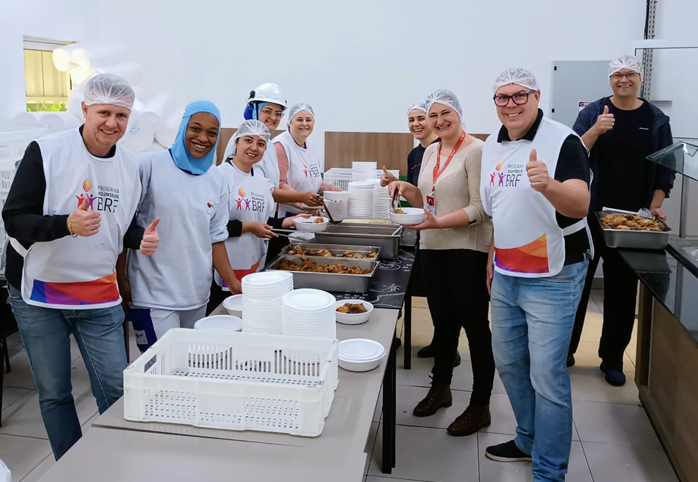 Foto de um grupo diverso de pessoas em uma cozinha montando refeições. Alguns estão com colete utilizado nas ações de voluntariado do Instituto BRF. Todos estão sorrindo.