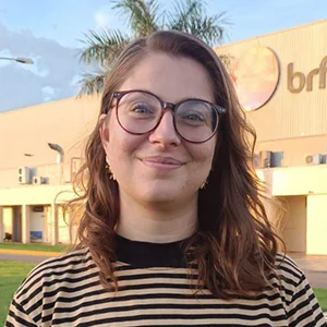 Foto de Gabriele Candido, uma mulher branca, com cabelos levemente ondulados na altura dos ombros. Ela está com óculos de grau com armação redonda, blusa listrada e sorri levemente.