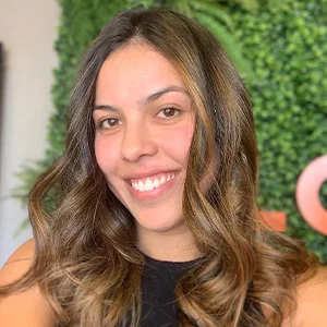 Foto de Isabela Ramos Esteves, uma mulher branca, com cabelos levemente ondulados. Ela usa uma blusa preta sem mangas e sorri.