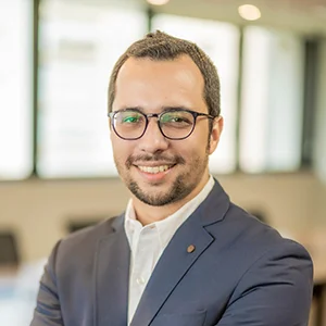 Foto de Pietro Carlos de Souza Rodrigues, um homem de pele branca, levemente calvo, com cabelos curtos e cavanhaque. Ele está de óculos de grau com armação preta, uma camisa social com um blazer por cima e sorri.