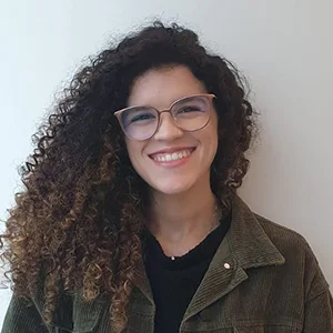 Foto de Rafaela Berçot, uma mulher branca, com cabelos cacheados volumosos. Ela está com óculos de grau, jaqueta marrom por cima de blusa preta e sorri.