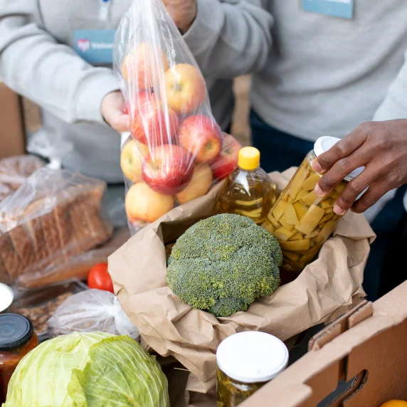 Foto de duas pessoas colocando alimentos em uma caixa. Uma delas segura um saco plástico com maçãs, enquanto a outra coloca um vidro de conserva em uma sacola de papelão, que já  contém óleo e um maço de brócolis.
