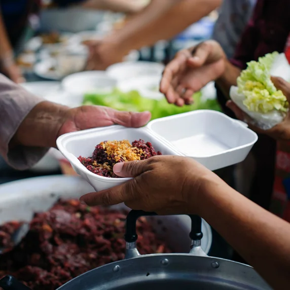 Foto das mãos de uma pessoa entregando embalagem de isopor com comida para outra pessoa.