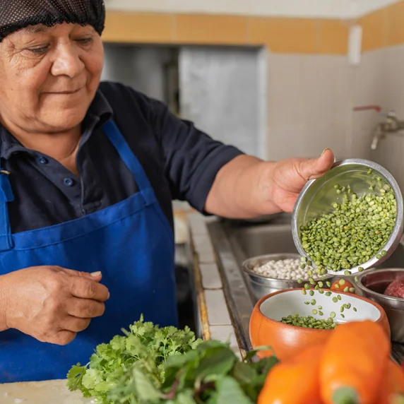 Foto de mulher em uma cozinha. Ela veste avental e touca que protege os cabelos, e despeja grãos de lentilha em uma tigela.